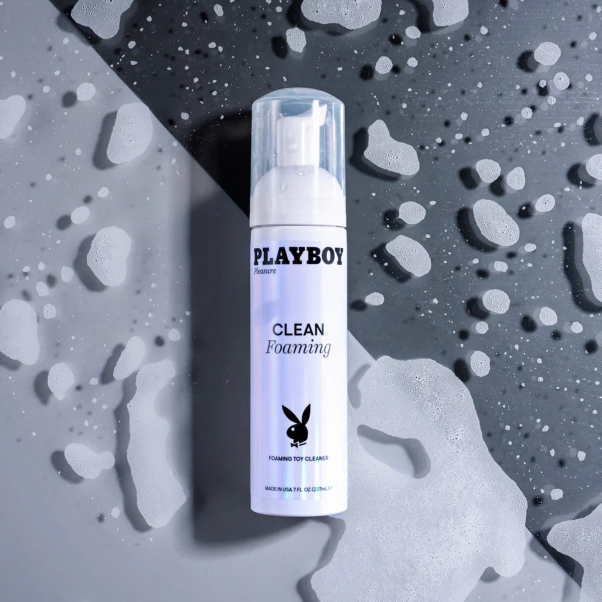 Playboy Pleasure Clean Foaming Toy Cleaner bottle lying flat on a grey, wet surface surrounded by soapy foam bubbles.