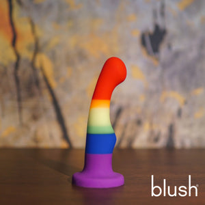 Side view of the blush Avant Pride Freedom Plug placed on its suction cup on wooden surface, with an autumn themed background. On the bottom right of the image is the blush logo