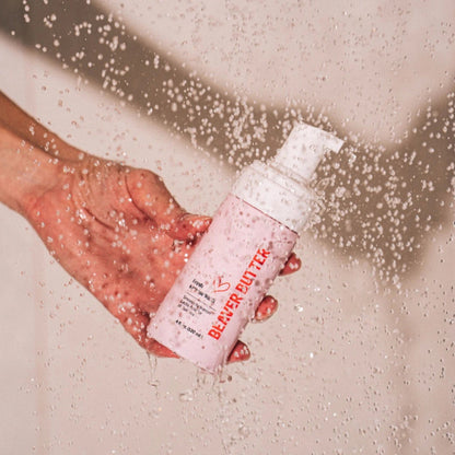 A hand holding the pink Beaver Butter Intimate Wash bottle in a shower setting with water droplets splashing around the pump.