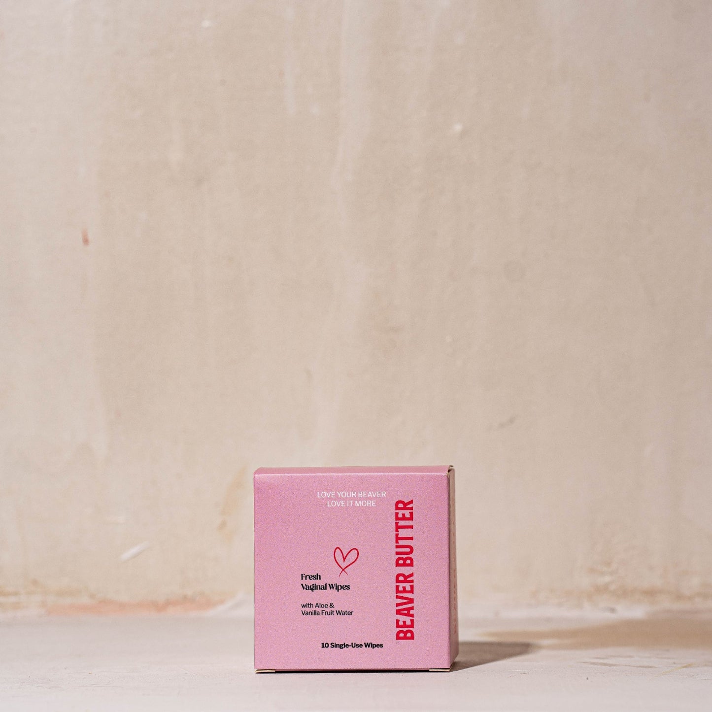 A minimalist product shot of the pink Beaver Butter Fresh Vaginal Wipes box containing 10 single-use wipes, styled upright on a rustic neutral surface against a clean studio background.