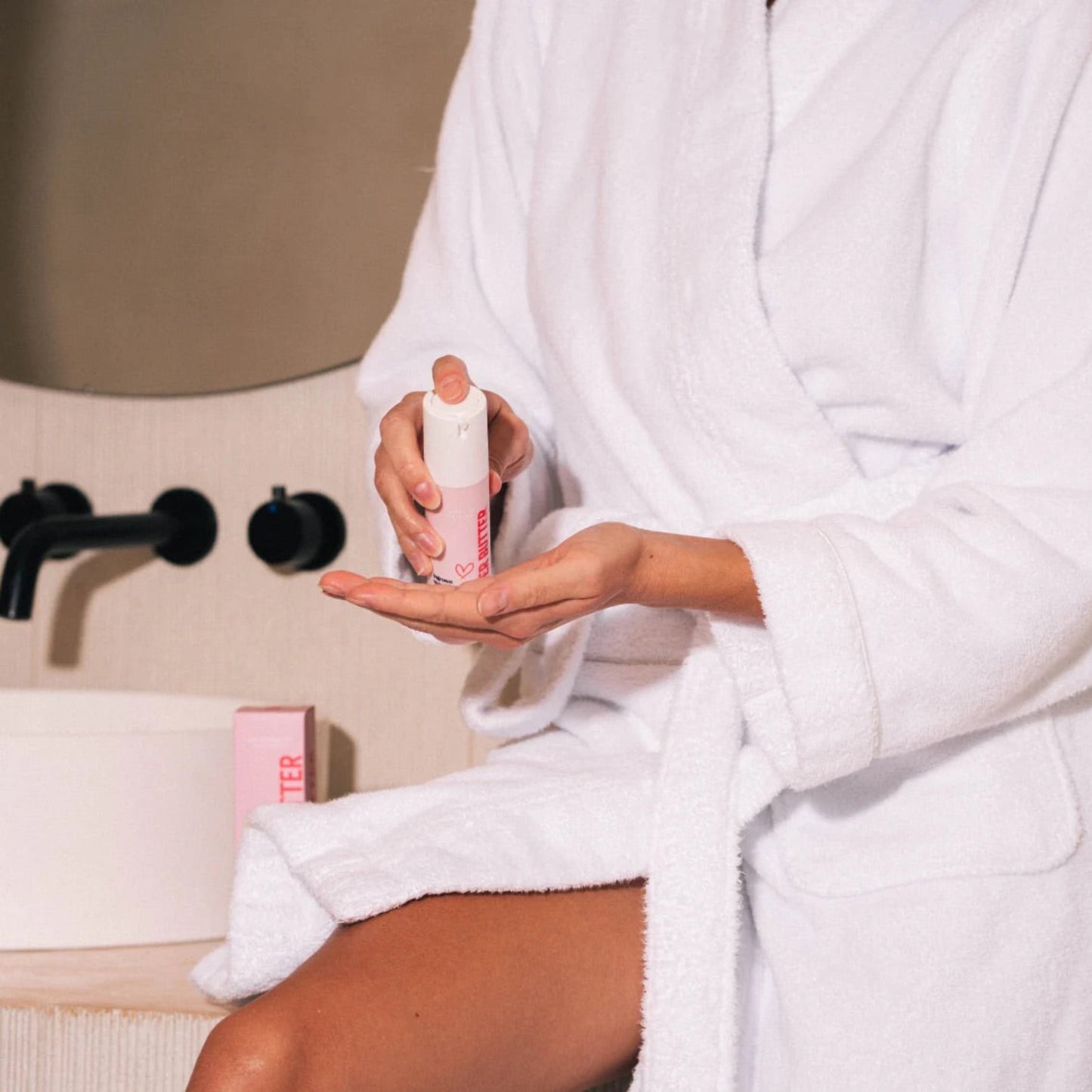 A woman in a white bathrobe sitting in a bathroom, applying Beaver Butter Ingrown Hair Serum to her leg to prevent razor bumps.