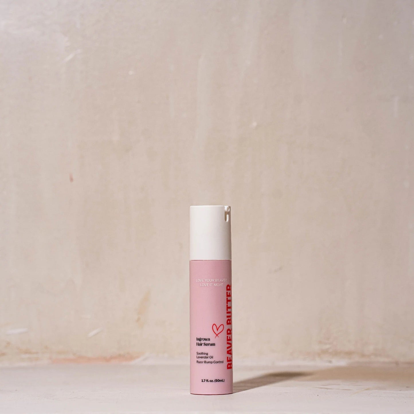 The pink Beaver Butter Ingrown Hair Serum bottle standing against a textured wall, showcasing the "Love Your Beaver" branding.