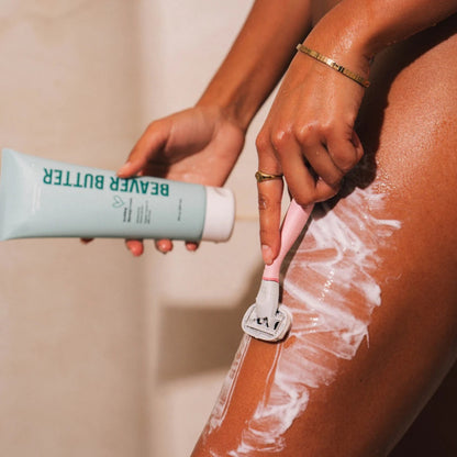 A person in a shower setting using a pink razor to shave their leg with Beaver Butter.