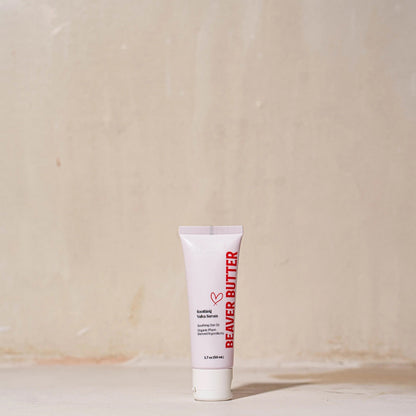 A studio shot of the Beaver Butter Soothing Vulva Serum tube standing upright on a neutral, textured surface with soft lighting.