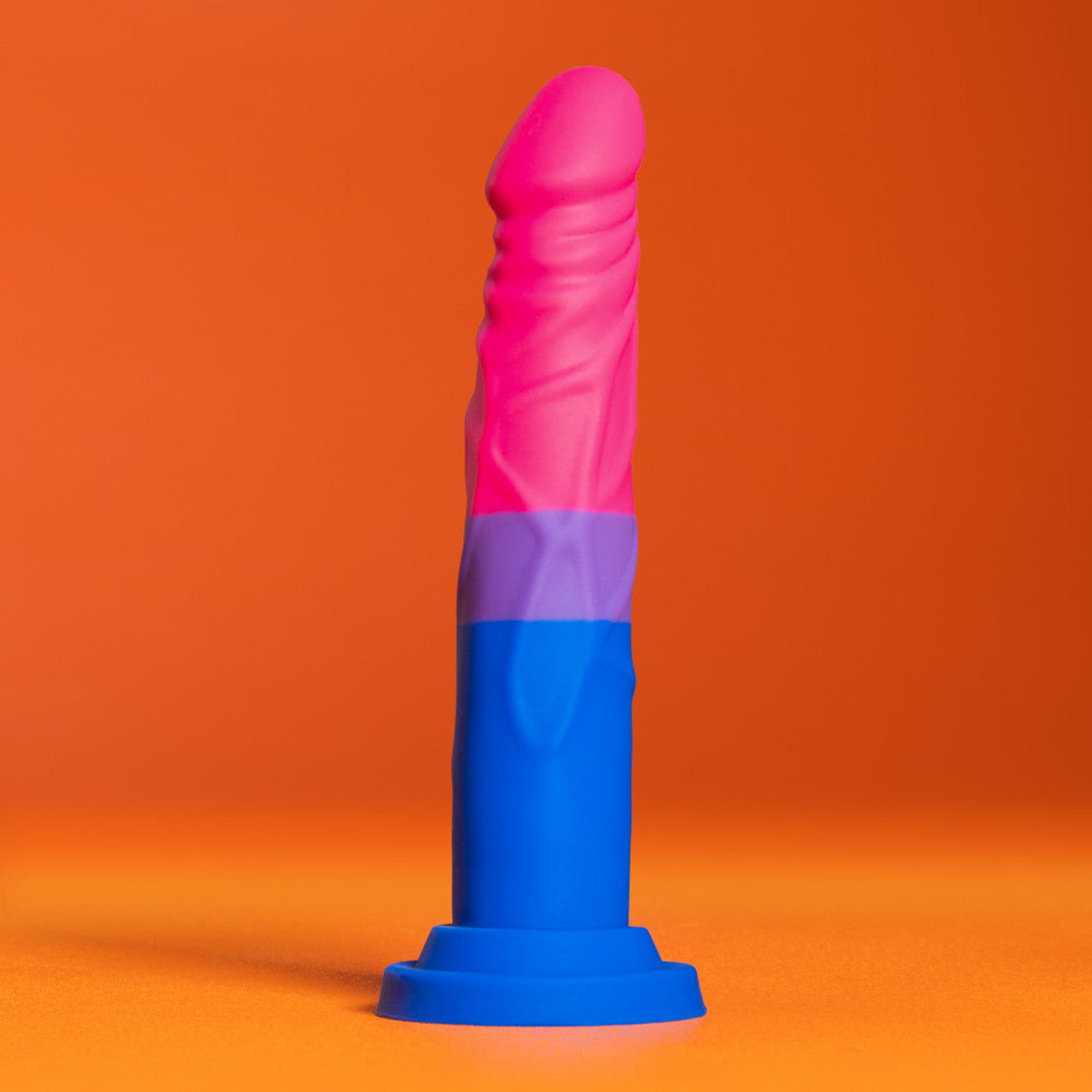 Side view of the blush Avant Pride Love Dildo, placed on its suction cup, with an orange background.