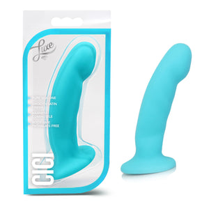 On the left side of the image is the product packaging. On the packaging is the Luxe logo, the product inside fully visible through clear packaging, product features: Pure silicone; Smooth satin finish; Harness compatible; Body safe phthalate free, and the product name: Cici. Beside the packaging is the product blush Luxe CiCi Dildo, stood up on its suction cup base.