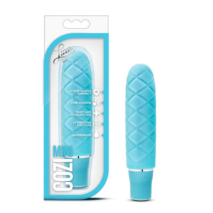 On the left side of the image is the aqua variant product packaging. On the packaging is the Luxe logo, product feature icons for: 5 year global warranty; Pure silicone; Body safe phthalate free; 10 vibrating functions; waterproof, product name: Cozi mini, and the product inside fully visible through clear packaging. Beside the packaging, is the product blush Luxe Cozi Mini Aqua Vibrator, standing up on its base.