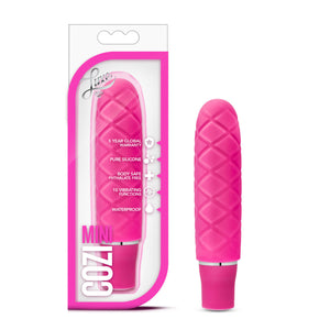 On the left side of the image is the faschia variant product packaging. On the packaging is the Luxe logo, product feature icons for: 5 year global warranty; Pure silicone; Body safe phthalate free; 10 vibrating functions; waterproof, product name: Cozi mini, and the product inside fully visible through clear packaging. Beside the packaging, is the product blush Luxe Cozi Mini Faschia Vibrator, standing up on its base.