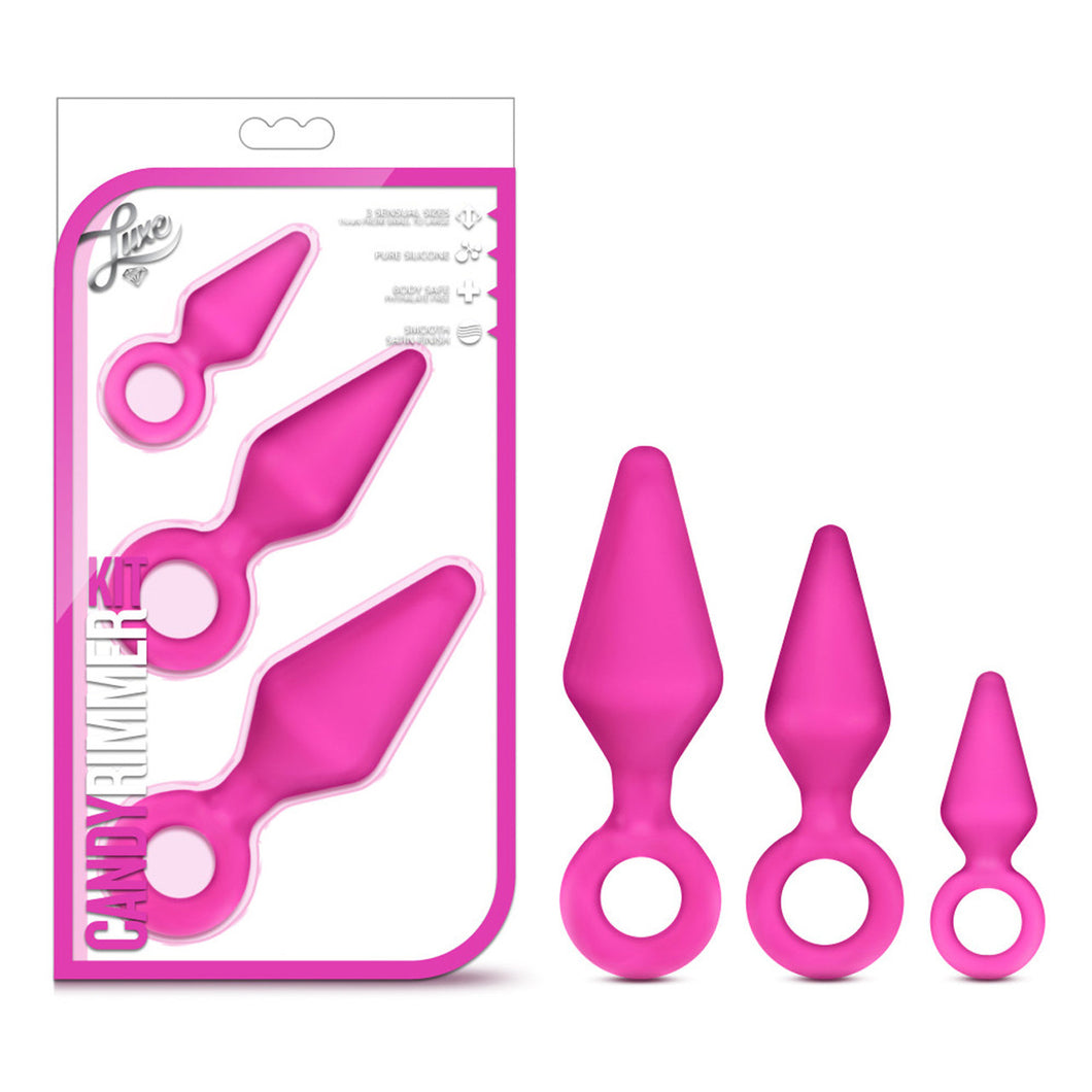 On the left side of the image is the packaging. On the packaging is the Luxe logo, product feature icons for: 3 sensual sizes; Pure silicone; Body safe; Smooth Satin finish, the product fully visible through clear packaging, and in the bottom left is product name: Candy Rimmer Kit. Beside the packaging is the product blush Luxe Candy Rimmer plugs stood up beside each other from Large to small.