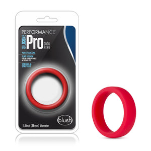 On the left side of the image is the packaging. On the packaging is the Performance logo, product name: Silicone Pro Cock Ring, product features: Pure silicone; Flat design for a comfortable & secure fit; Strong & stretchy, in the middle is the product inside visible through clear packaging, at the bottom "1.5inch (38mm) diameter" and the blush logo in the right corner. Beside the packaging is the blush Performance Silicone Pro red Cock Ring.