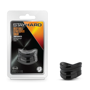 On the left side of the image is the product packaging. On the packaging is the Stay Hard logo, product name: Beef Ball Stretcher Snug 1 Inch Diameter, product features: Superior Strength; Body Safe; Elastic, the product inside visible through clear packaging, blush logo, slogan: Perform like a stud, and an icon for Body safe - Laboratory certified. Beside the packaging is the product blush Stay Hard Beef Ball Stretcher Snug - 1 Inch Diameter.