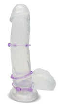Load image into Gallery viewer, Side view of a clear dildo standing on its suction cup base, with 3 blush Stay Hard Beaded Cock Rings wrapped around the middle of the shaft, at the base of the shaft, and at the base of the dildo, above the suction cup. Showing the placements of the Cock Rings.