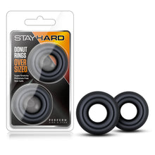 Load image into Gallery viewer, On the left side of the image is the product packaging. On the packaging is the Stay HArd logo, product name: Donut Rings Oversized, product features: Super stretchy; phthalate free; Skin safe, the product inside visble through clear packaging, and the slogan on bottm right: Perform like a stud. Beside the packaging is the product blush Stay Hard Donut Rings Oversized with the left standing slightly infront of the other.