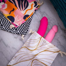 Load image into Gallery viewer, On the top left of the image are 2 style variants of the blush The Collection Cotton Toy Bags, laying on top is the Burst style variance, and on the bottom is the Bomba style variant. From the lower middle of the image is the Embrace variant toy bag, and from the inside sticking out from the bag are two sex toys.