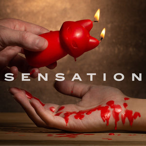 An image of a male's hand holding a lit up blush Temptasia Fox Drip Candle, and dripping its wax on top of a female's palm. In the middle is a caption text: Sensation.