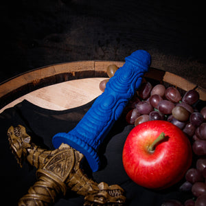 The blush The Realm Draken Snap On Dildo is snapped on the Drago Lock On Sword Handle, and laying flat on top of a bushel of grapes, beside an apple.