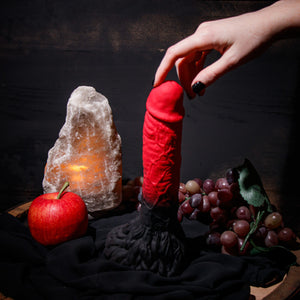 Side view of the blush The Realm Lycan Lock On Dildo, standing on its base beside an Apple, and a bushel of grapes, with a female's hand reaching down, and gently touching the tip of the dildo.