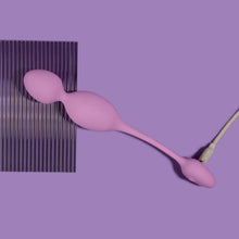 Charger l&#39;image dans la galerie, blush Wellness Raine Vibrating Kegel Ball stretched out and laying flat over a plastic material, without the wireless remote control, and plugged in from the bottom, showing the charging port.
