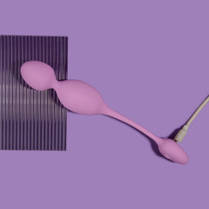 blush Wellness Raine Vibrating Kegel Ball stretched out and laying flat over a plastic material, without the wireless remote control, and plugged in from the bottom, showing the charging port.