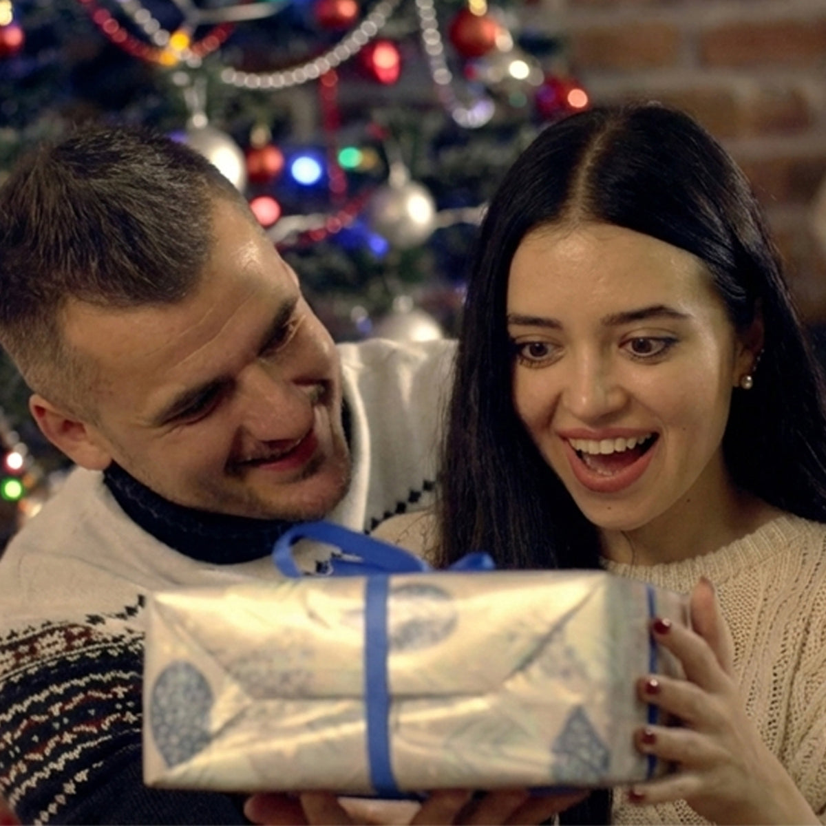 A man gifting a present to a woman in front of a decorated Christmas tree.