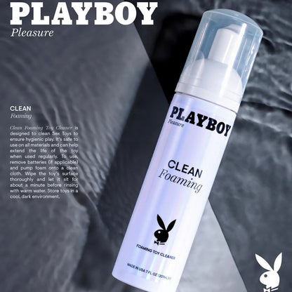 Angled view of the Playboy Pleasure Clean Foaming Toy Cleaner bottle resting on a dark, wet, textured surface.