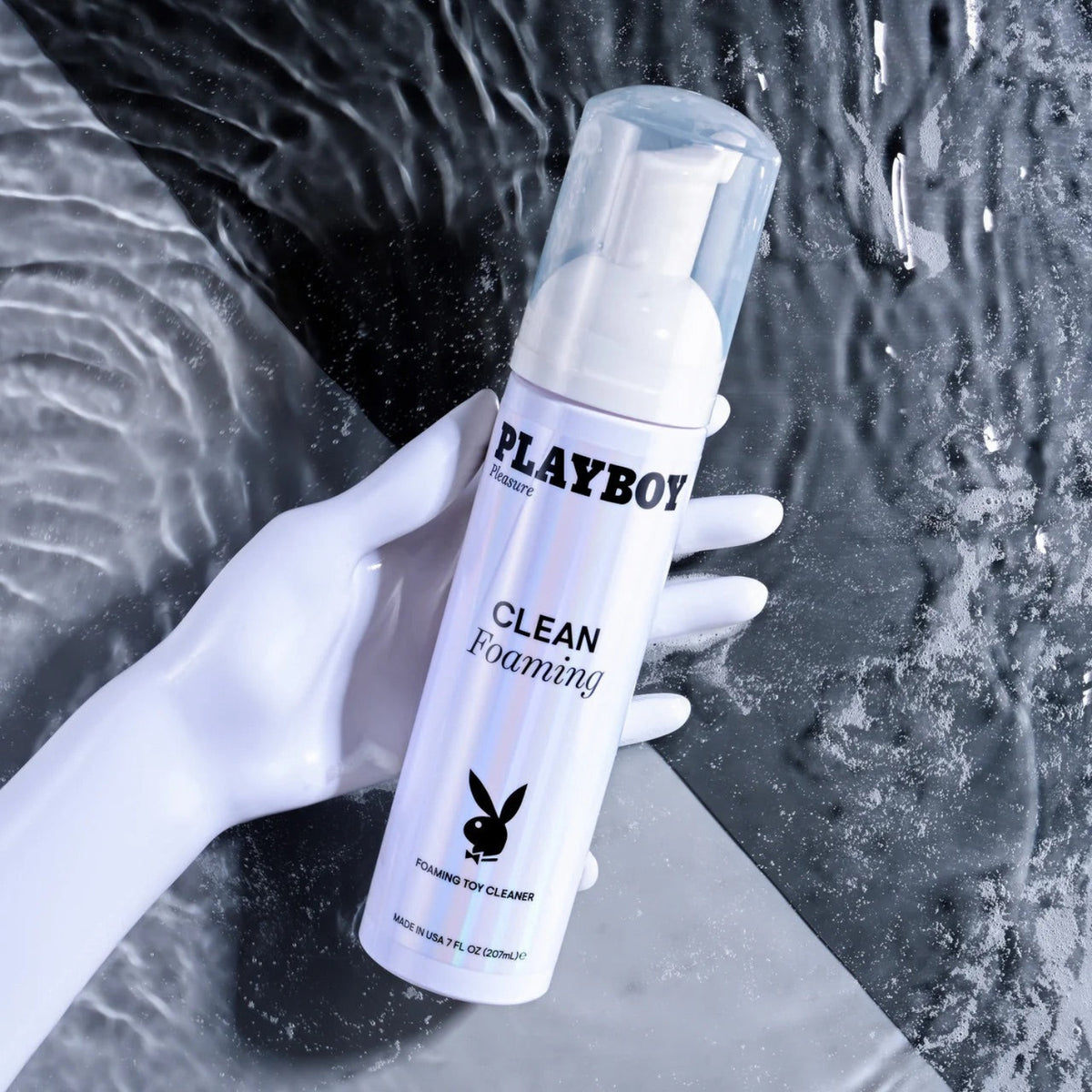 A white mannequin hand gripping a bottle of Playboy Pleasure Clean Foaming Toy Cleaner against a dark, rippling water background.