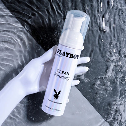 A white mannequin hand gripping a bottle of Playboy Pleasure Clean Foaming Toy Cleaner against a dark, rippling water background.