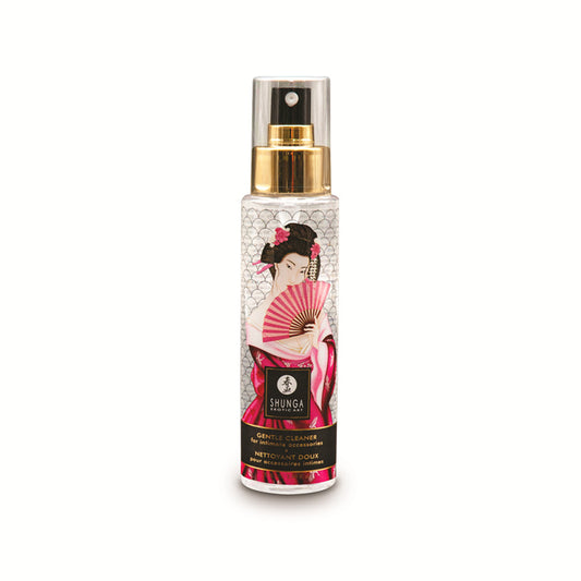 A tall, clear spray bottle of Shunga Gentle Cleaner for Intimate Accessories, featuring gold-tone hardware and a decorative label with an artistic illustration of a woman in a pink kimono holding a folding fan.