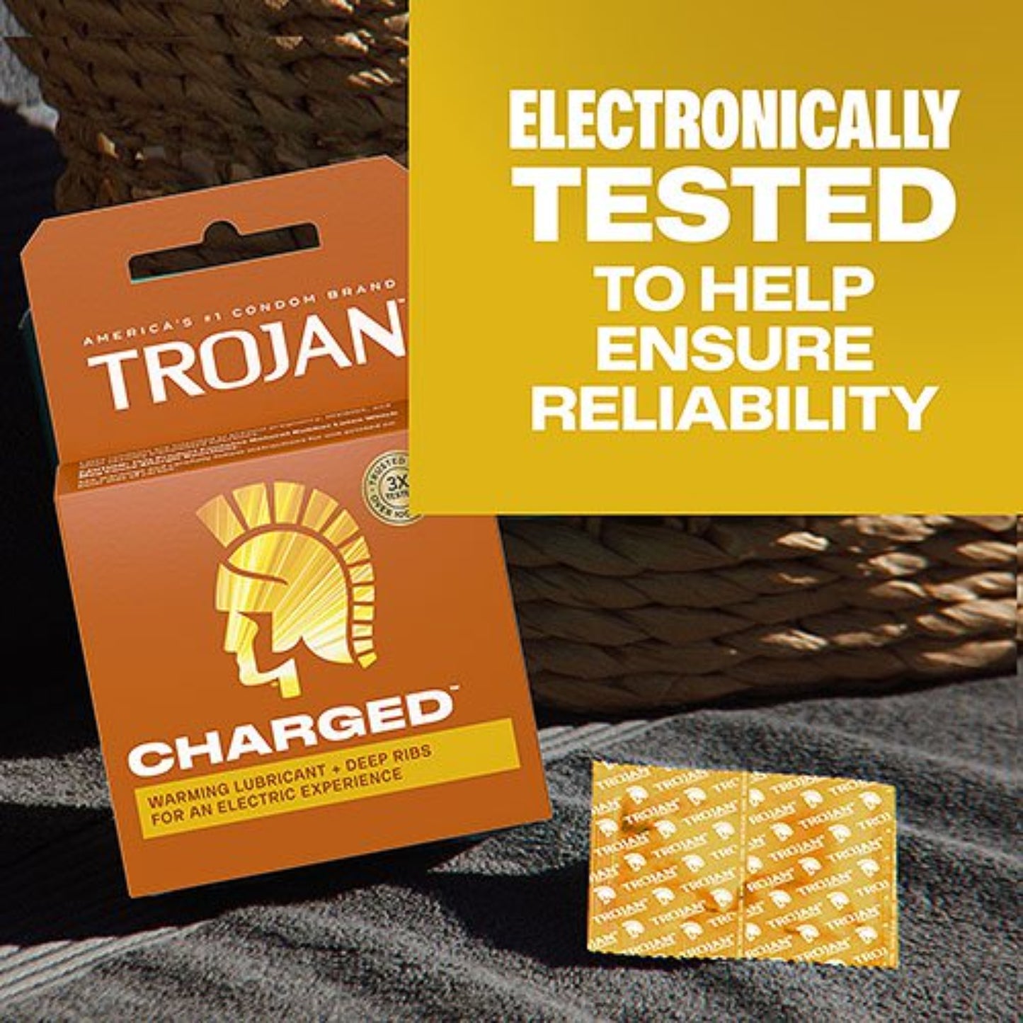 A box of Trojan Charged Warming Lubricant and Deep Ribs condoms resting on a dark grey towel next to an individually wrapped foil condom wrapper, featuring bold text reading "Electronically tested to help ensure reliability."